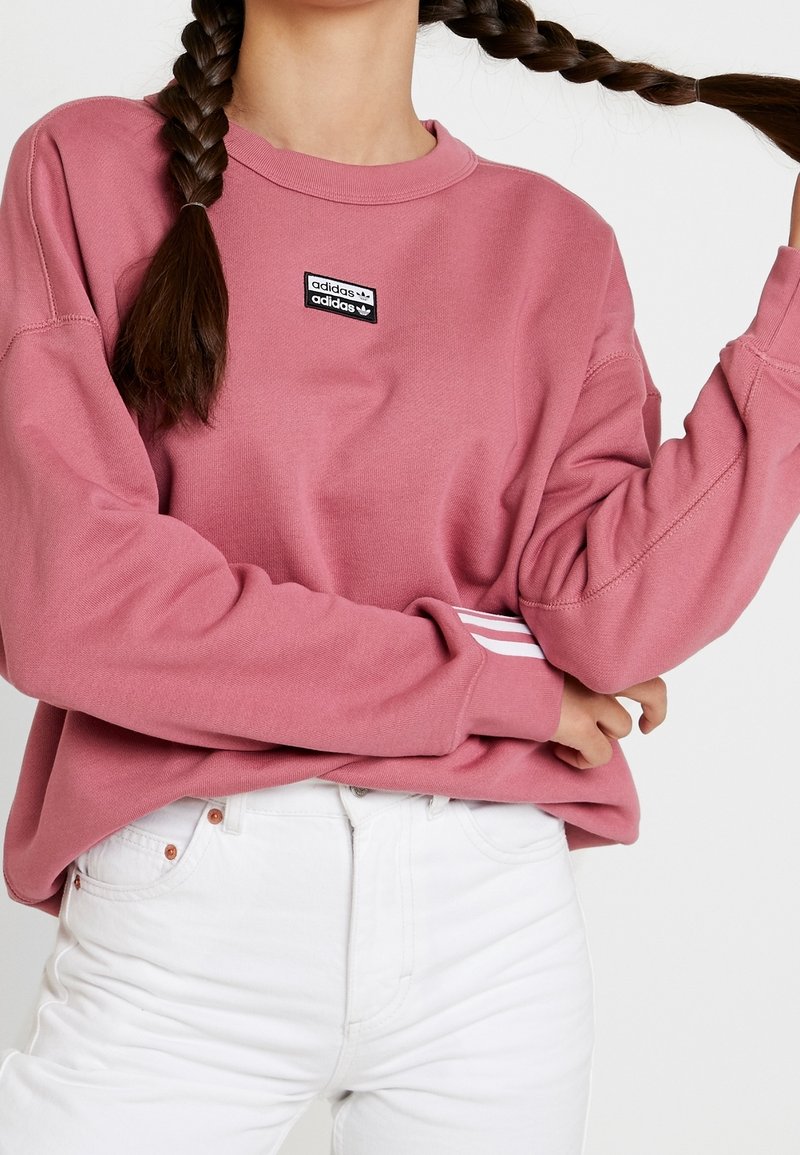 Sweatshirt rose avec des manches longues, col côtelé et patch logo Adidas noir sur la poitrine. Présente des rayures blanches sur les poignets. Porté avec un pantalon blanc.