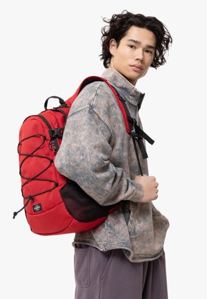 Zaino rosso con una finitura texturizzata, spallacci regolabili, corda elastica bungee sul davanti e tasche laterali in rete. Indossato da una persona in abbigliamento informale.