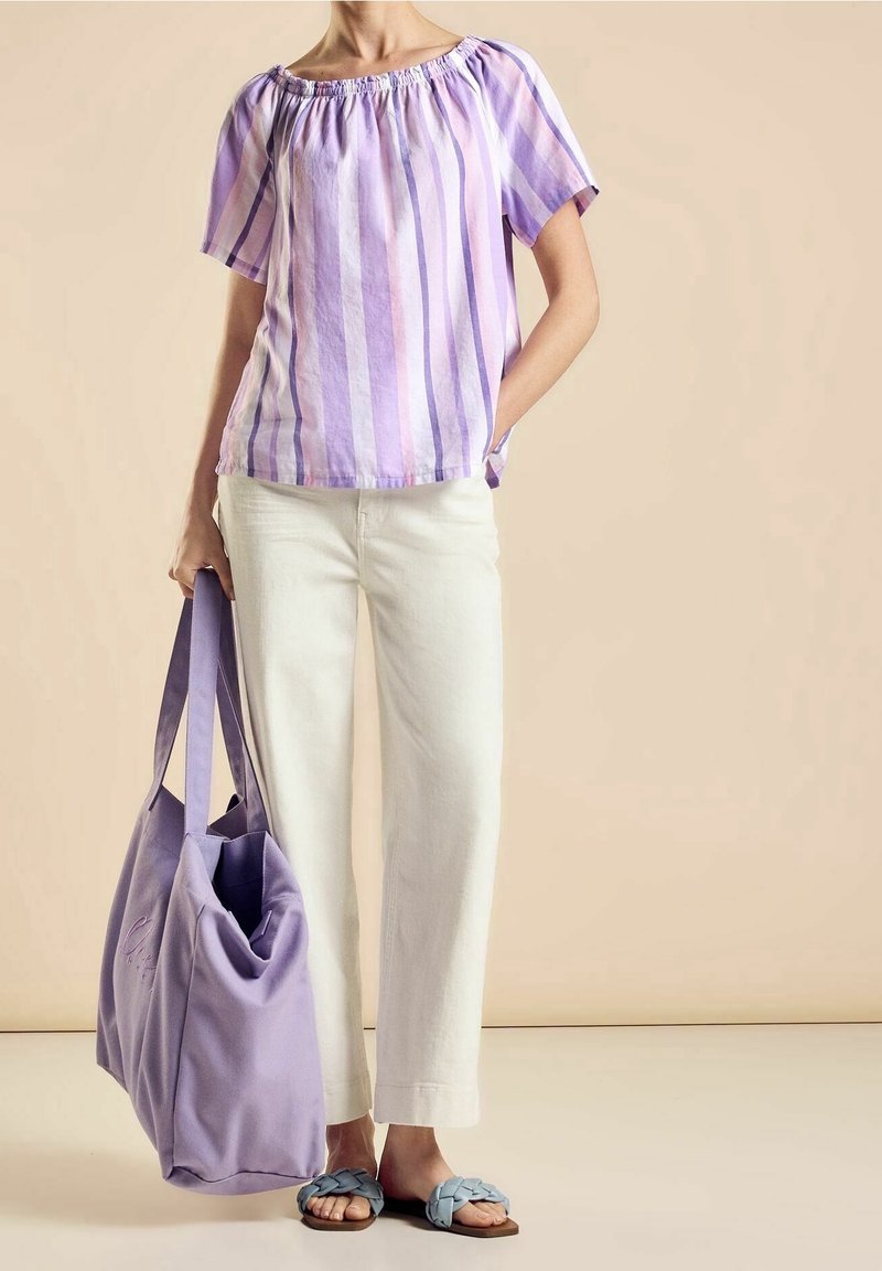 Personne portant un blouse rayée lavande et blanche et un pantalon blanc, tenant un grand sac fourre-tout lavande et portant des sandales à brides bleues.