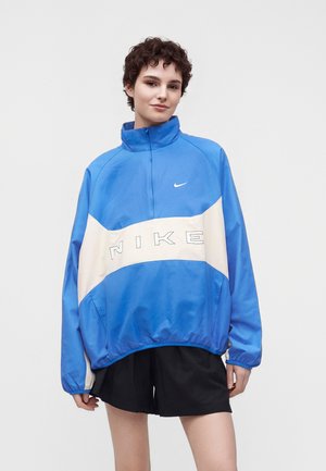 Jeune personne portant une veste coupe-vent Nike bleue avec une bande horizontale blanche et un short noir, debout devant un fond uni.