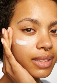 Feuchtigkeitsspendende Creme wird auf die Wange einer Person mit mittelbrauner Haut aufgetragen, die einen runden Septumring und strukturierte lockige Haare hat.