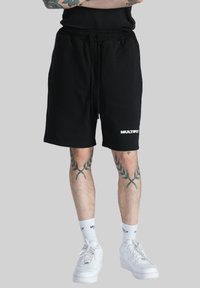 Pantaloni corti neri in cotone con una cintura elastica, dotati di un logo bianco sul lato inferiore. Indossati con sneakers bianche e calzini.