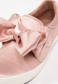 Baskets en satin rose avec un large nœud à l'avant, bout rond et semelle en caoutchouc blanche. La surface a une texture lisse et brillante.