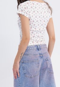 Mujer joven que lleva una blusa blanca de flores de manga corta y unos pantalones cortos de mezclilla azul de cintura alta, mostrando un tatuaje en su antebrazo izquierdo.