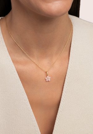 Goldene Halskette mit einem rosa Blumenanhänger und drei kleinen weißen Steinen in der Mitte. Der Hintergrund zeigt einen strukturierten, hellen Stoff.
