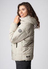 Beige donsjas met een capuchon, voorzien van een doorgestikt ontwerp, een logopatch op de mouw en zijzakken. Gemaakt van glad, geïsoleerd materiaal.