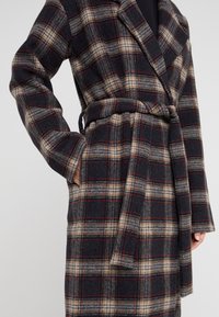 Abrigo de mezcla de lana con estampado de cuadros en gris oscuro, beige y rojo. Presenta un cuello de solapa, cintura con cinturón y bolsillos laterales. Textura suave con un diseño estructurado.