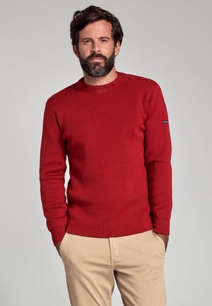 Czerwony sweter z prążkowanego dzianiny z okrągłym dekoltem, będący ozdobiony czterema dekoracyjnymi guzikami na ramieniu oraz subtelnym logo marki na rękawie.