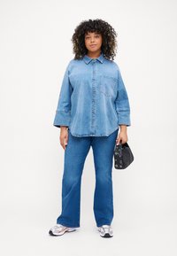 Camisa de ganga azul com design abotoado, mangas longas e bolso no peito. Combinada com jeans azuis de boca de sino e mala preta.