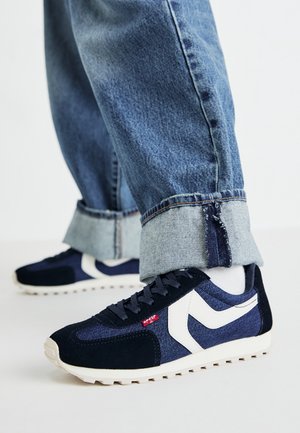 Zapatillas navy con una mezcla de ante y malla, que presentan detalles en blanco, un patrón texturizado y una suela de goma. Combinadas con unos jeans de denim acampanados.