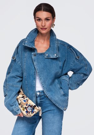 Femme portant une veste en jean bleue oversize et un jean, tenant un petit sac à main tissé à motif floral sur un fond uni.