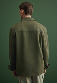 Chaqueta texturizada de color verde olivo con cuello, mangas largas y puños abotonados, vista desde atrás sobre un fondo verde oscuro.