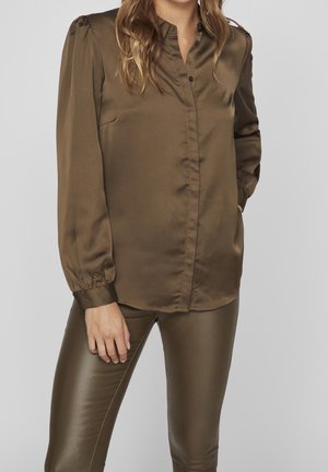 Femme portant un chemisier en satin vert olive boutonné avec des manches bouffantes et un pantalon assorti effet cuir, debout, une main dans la poche.