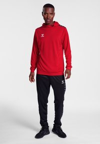 Rød langærmet sportstrøje med hætte og logo, parret med sorte atletiske bukser med sidelister og en tætsiddende pasform.