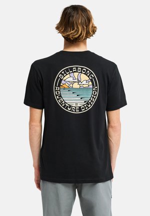 Personne aux cheveux courts et bruns portant un t-shirt noir avec un logo rond coloré Billabong Adventure Division montrant le soleil, les nuages et les vagues de l’océan dans le dos.