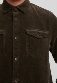 Chemise marron en velours côtelé avec un col pointu, une fermeture à boutons sur le devant et deux poches poitrine. Le tissu présente une texture côtelée.