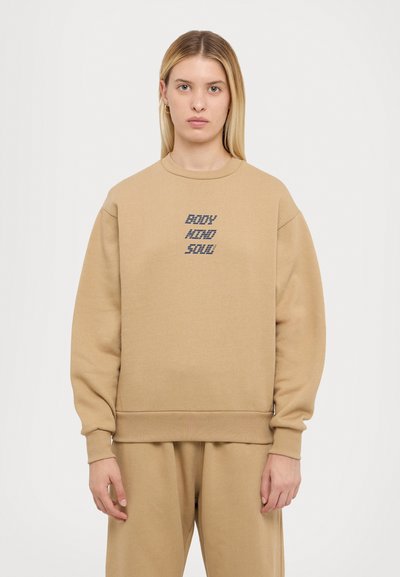 Beige sweatshirt with a crew neckline, long sleeves, and ribbed cuffs. Features "BODY MIND SOUL" text in blue. Soft, smooth material.