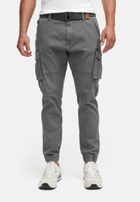 Homme portant un pantalon cargo gris avec des bas élastiques, une ceinture noire, des baskets blanches et une chemise blanche sur un fond blanc uni.