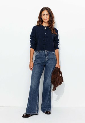 Cardigan côtelé marine avec des boutons dorés, associé à un jean flare taille haute. Sac en daim marron avec franges. Des chaussures lisses et brillantes complètent le look.