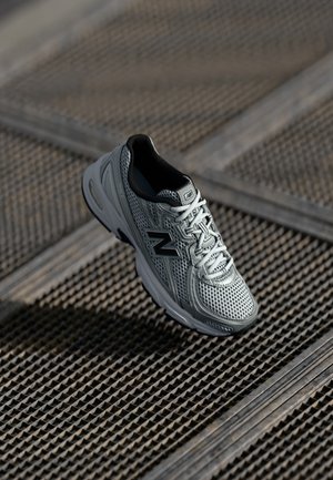 Baskets New Balance grises et noires avec un design en mesh reposant sur une surface de grille métallique texturée.