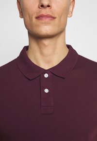 Bordeaux polo shirt met kraag, gemaakt van katoen, met drie witte knopen en een gladde textuur. Eenvoudig, klassiek ontwerp.