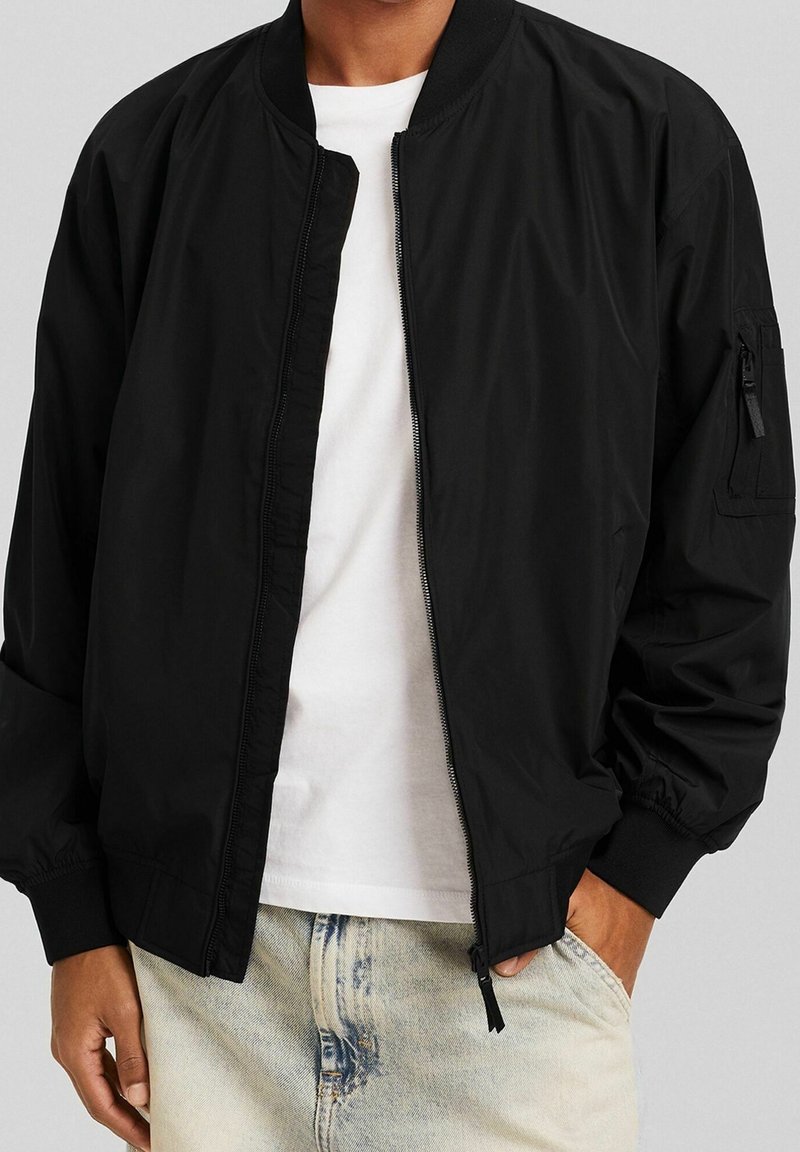 Veste aviateur noire en matériau léger, dotée d'une fermeture éclair à l'avant, de poignet côtelé et d'une poche utilitaire sur la manche gauche.