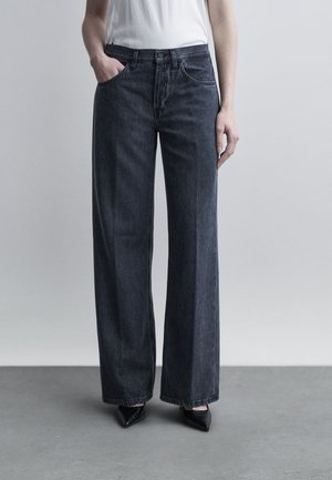 Άτομο που φοράει τζιν με ψηλή μέση και φαρδιά παντελόνια σε σκούρο χρώμα, με το ένα χέρι στην τσέπη και μαύρα παπούτσια με μυτερή μύτη σε γκρι πάτωμα.