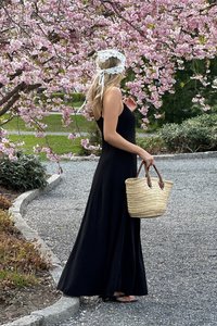 Svart maxiklänning med en flödande silhuett, kombinerad med en halm- och läderhandväska i brunt, placerad mot rosa körsbärsblommor.