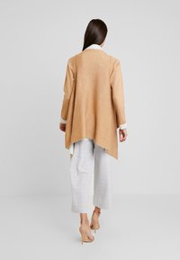 Beige langer Cardigan mit asymmetrischem Saum, kombiniert mit hellen karierten Hosen. Das Outfit verfügt über einen cremefarbenen Kragen und neutrale Pumps.