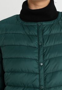 Veste puffer verte en matériau matelassé avec un col montant, dotée de boutons pression à l'avant et d'une texture lisse et brillante.