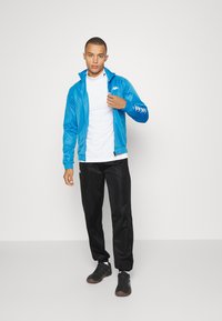 Muž oblečený v modrej zapínacej bundě, bielom roláku, čiernych teplákach a čiernych teniskách stojí na bielom pozadí.