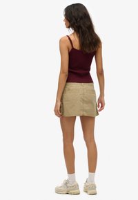 Kobieta o długich ciemnych włosach, ubrana w bordo tank top, beżową mini spódnicę typu cargo, białe skarpetki i beżowe sneakersy, stojąca tyłem.