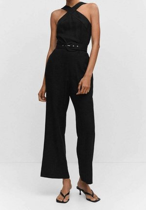 Femme portant une combinaison noire sans manches avec un col halter croisé et une ceinture, associée à des sandales à talons noires ouvertes à l’avant.