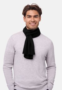 Écharpe en maille noire drapée autour du cou d'un homme, contrastant avec un pull gris clair. L'écharpe a une texture douce et des extrémités flottantes.