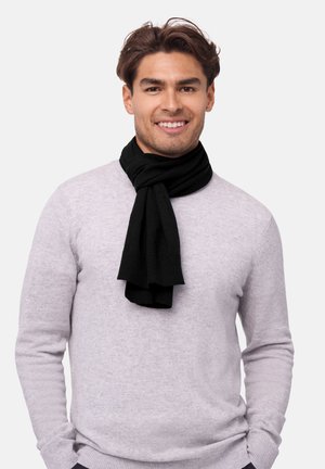 En sort strikket tørklæde hænger rundt om en mands hals og kontrasterer med en lysegul sweater. Tørklædet har en blød tekstur og løse ender.
