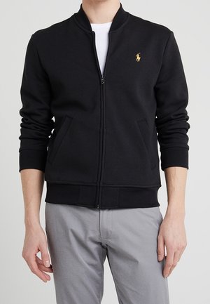 Mann trägt eine schwarze Jacke mit Reißverschluss und langen Ärmeln sowie ein kleines gelbes gesticktes Logo auf der linken Brust, kombiniert mit hellgrauen Hosen.