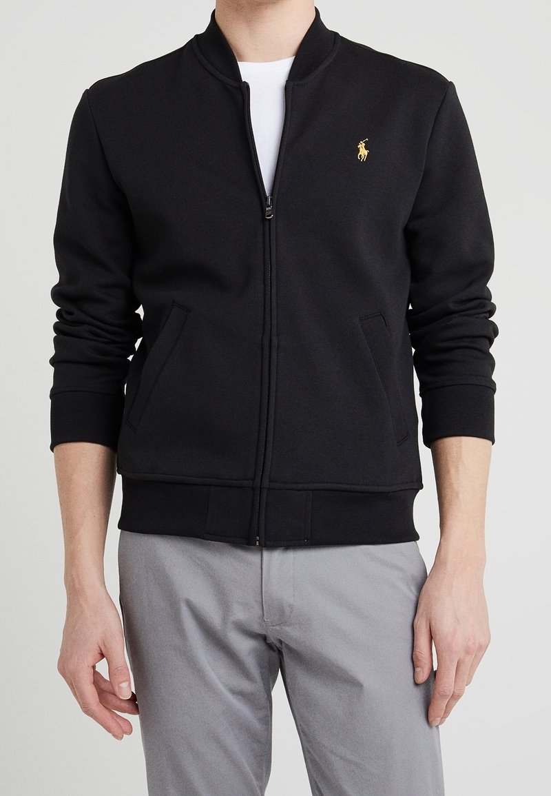 Homme portant une veste noire zippée à manches longues avec un petit logo brodé jaune sur la poitrine gauche, associée à un pantalon gris clair.