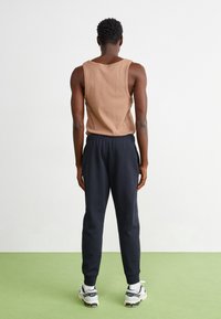 Brun linne med breda axelband, tillsammans med mörka joggerbyxor och vita sneakers, sedd bakifrån mot en ljus bakgrund.