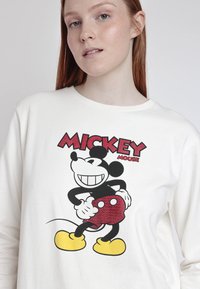 Camisa blanca de manga larga con un gráfico de Mickey Mouse en negro y rojo, con zapatos amarillos y un diseño redondo en pantalones cortos rojos.