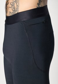 Close-up of black athletic leggings with zippered side pocket worn by tattooed person’s waist and hip visible.