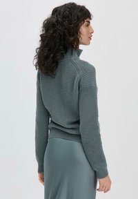Pull en laine gris avec col roulé, à bords côtelés, porté sur une jupe lisse de couleur vert argenté. La texture présente un motif épais.