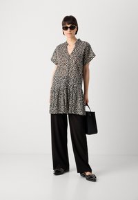 Kortermet tunika i svart-hvitt leopardmønster med V-hals og lagdelt kant, kombinert med svarte vide bukser og svarte loafers.