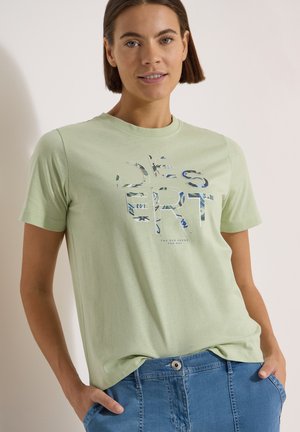 Femme aux cheveux bruns courts porte un t-shirt vert clair avec le texte "DESERT" et un motif floral, associé à un jean bleu, mains dans les poches.