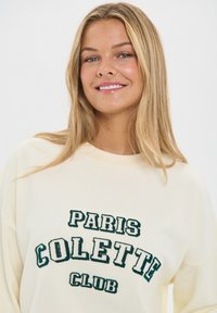 Krém színű pulóver laza szabásban, zöld hímzett felirattal: "PARIS COLETTE CLUB." Puha tapintás és laza kialakítás.