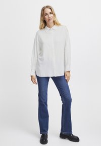 Camicia bianca con bottoni, maniche lunghe e vestibilità rilassata; abbinata a jeans svasati blu scuro; la modella indossa stivaletti neri alla caviglia.