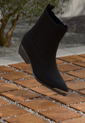 Musta nilkkuri, jossa on tekstuuriset sivut, terävä varvas ja matala korko. Korostaa kultakuvioista yksityiskohtaa, jossa lukee "GUESS" korossa. Asetettu kuvioidulle laattapinnalle.