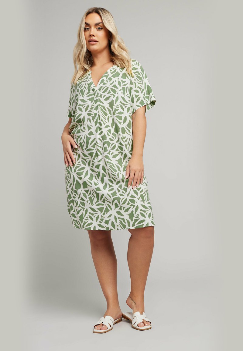 Robe verte à motifs avec des manches courtes, arborant un design floral blanc. Tissu léger, coupe décontractée, longueur mi-genou. Associée à des sandales blanches.