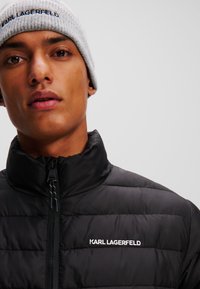 Zwarte pufferjas met hoge kraag, voorzien van een getextureerde quilt en een ritssluiting aan de voorkant. Grijze gebreide beanie met een logo-band.