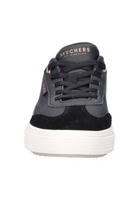 Zwarte Skechers sneaker met witte zool, zwarte veters en een gouden logo op de tong en zijkant, weergegeven vanuit de voorkant.