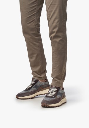 Benen in slim fit bruine broek met grijze, bruine en beige gestructureerde sneakers met witte veters, staand op een witte achtergrond.